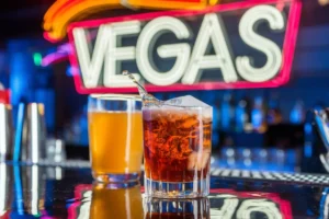 Vegas Bomb cocktail in a shot glass with a glass of energy drink on a glossy bar counter under neon lights.