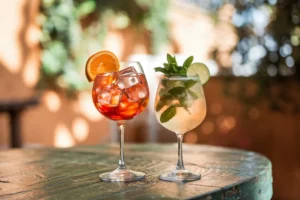 Realistic Aperol Spritz and Hugo Spritz served on a wooden table with orange and mint garnishes in a sunny Italian setting.